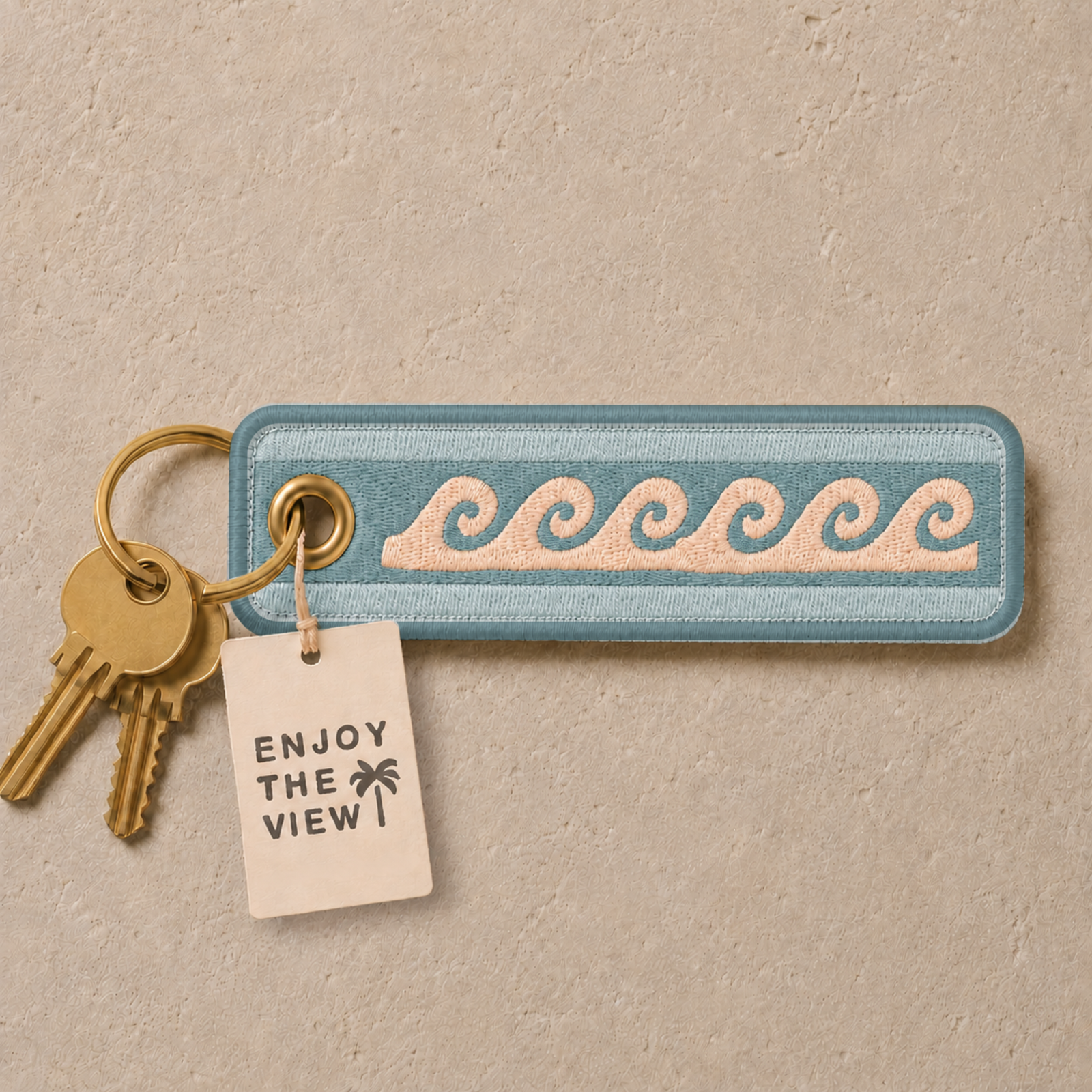 A rectangular embroidered patch with a light blue border and pale pink wave pattern, attached to a gold keyring with two keys and a small cream paper tag that reads “ENJOY THE VIEW” with a palm tree icon, on a textured beige background.