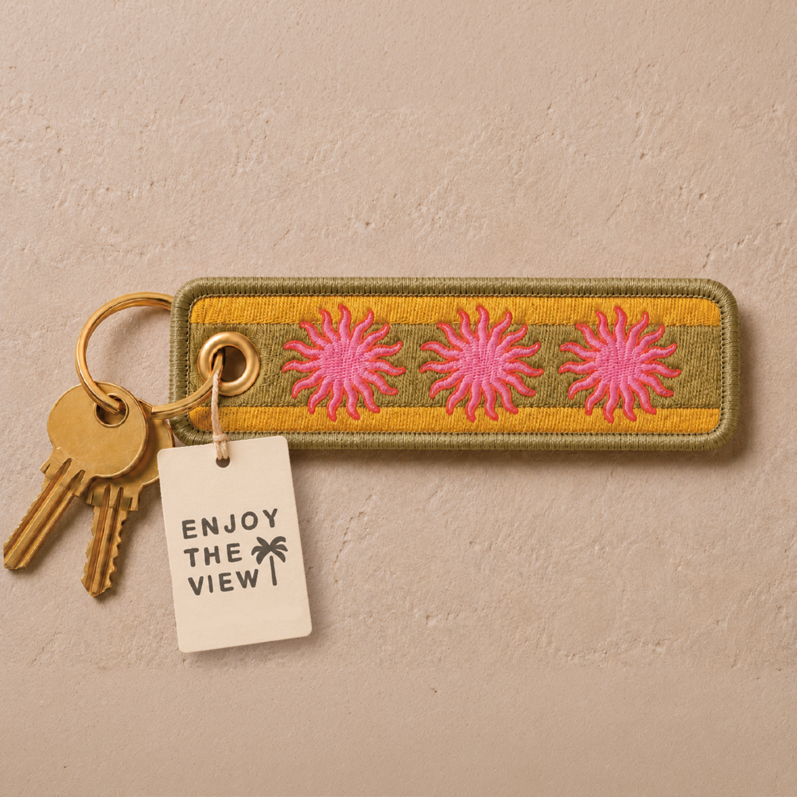 Embroidered rectangular keychain featuring three pink sunflower motifs on a yellow and olive green striped background, attached to a gold keyring with two keys and a white paper tag that reads 'ENJOY THE VIEW!'.