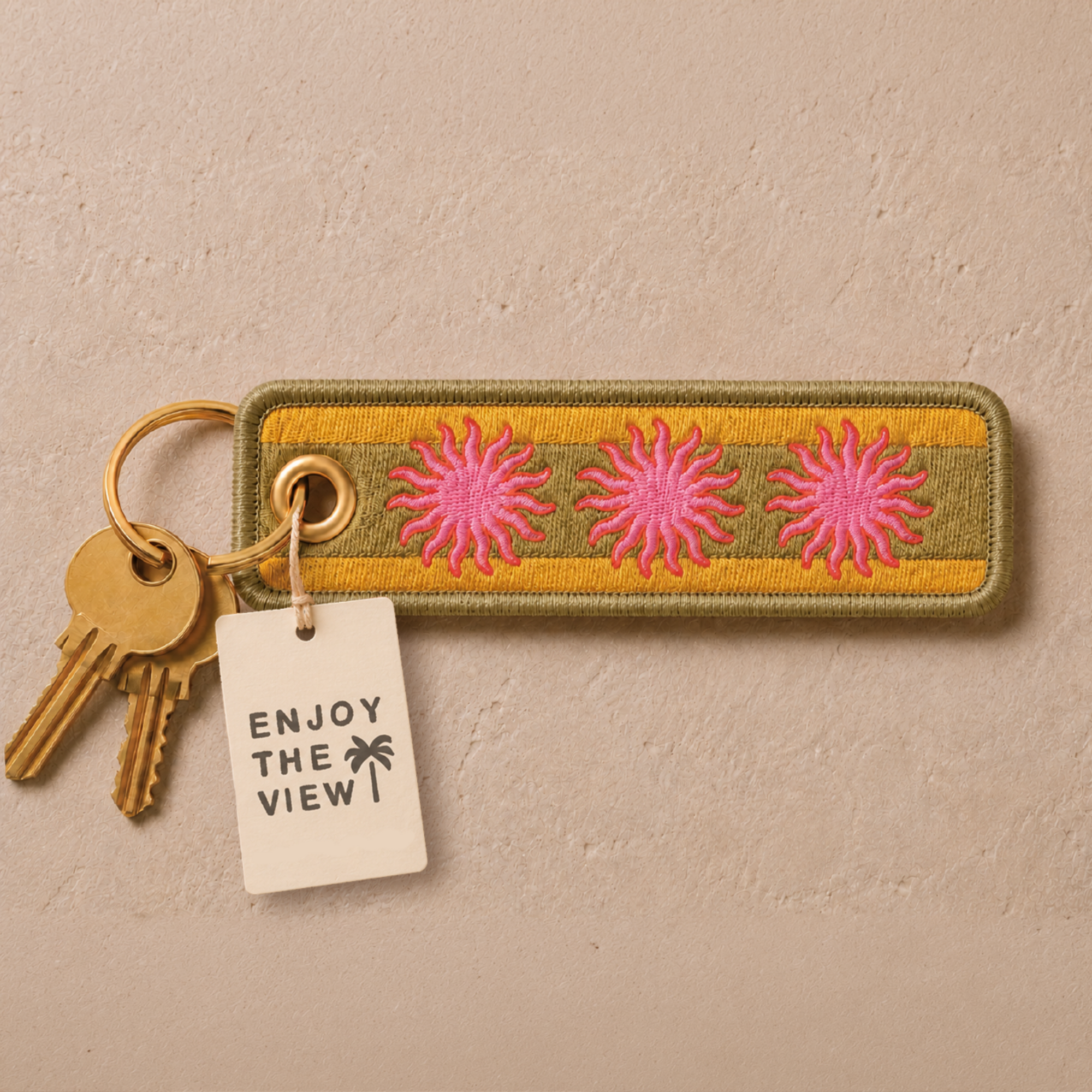 Embroidered rectangular keychain featuring three pink sunflower motifs on a yellow and olive green striped background, attached to a gold keyring with two keys and a white paper tag that reads 'ENJOY THE VIEW!'.