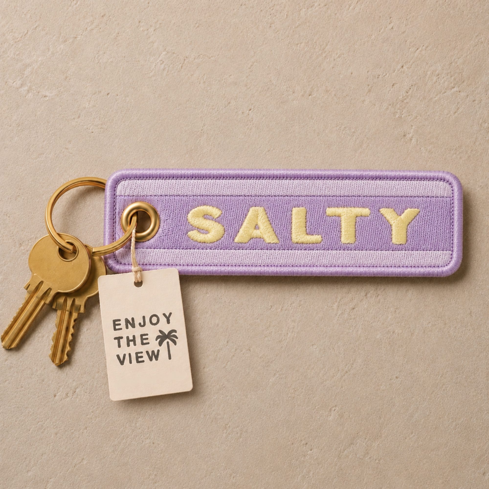 A lavender embroidered luggage tag with the word 'SALTY' in cream lettering, attached to a gold keyring with two gold keys and a small cream paper tag that reads 'ENJOY THE VIEW!'.