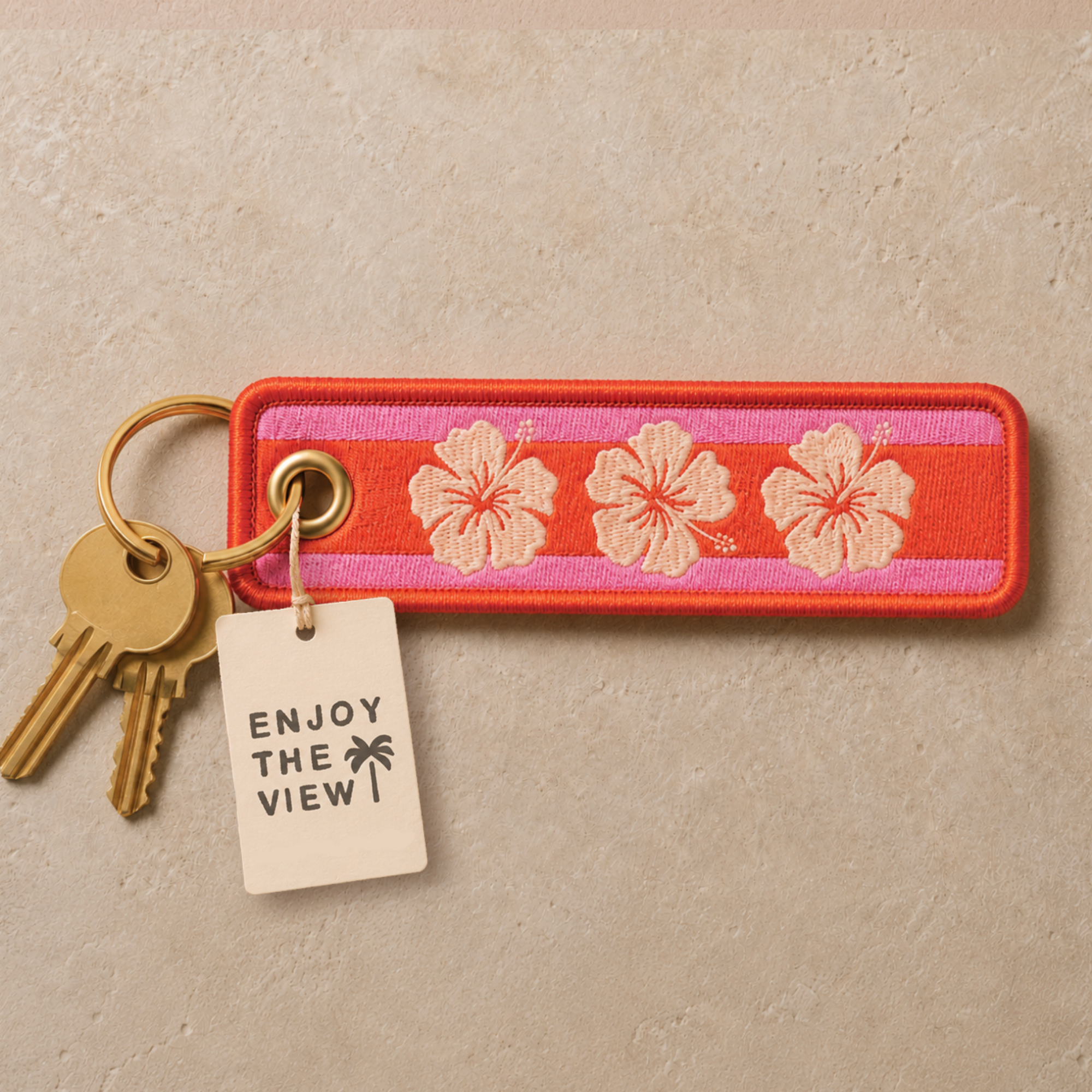 A rectangular embroidered keychain or luggage tag with three cream hibiscus flowers on a pink and orange striped background, attached to a gold key ring with two gold keys and a cream paper tag that reads 'ENJOY THE VIEW!'. The item is photographed on a textured beige stone surface.