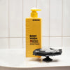 Yellow bottle of body wash with a black scrubber on a white tiled background