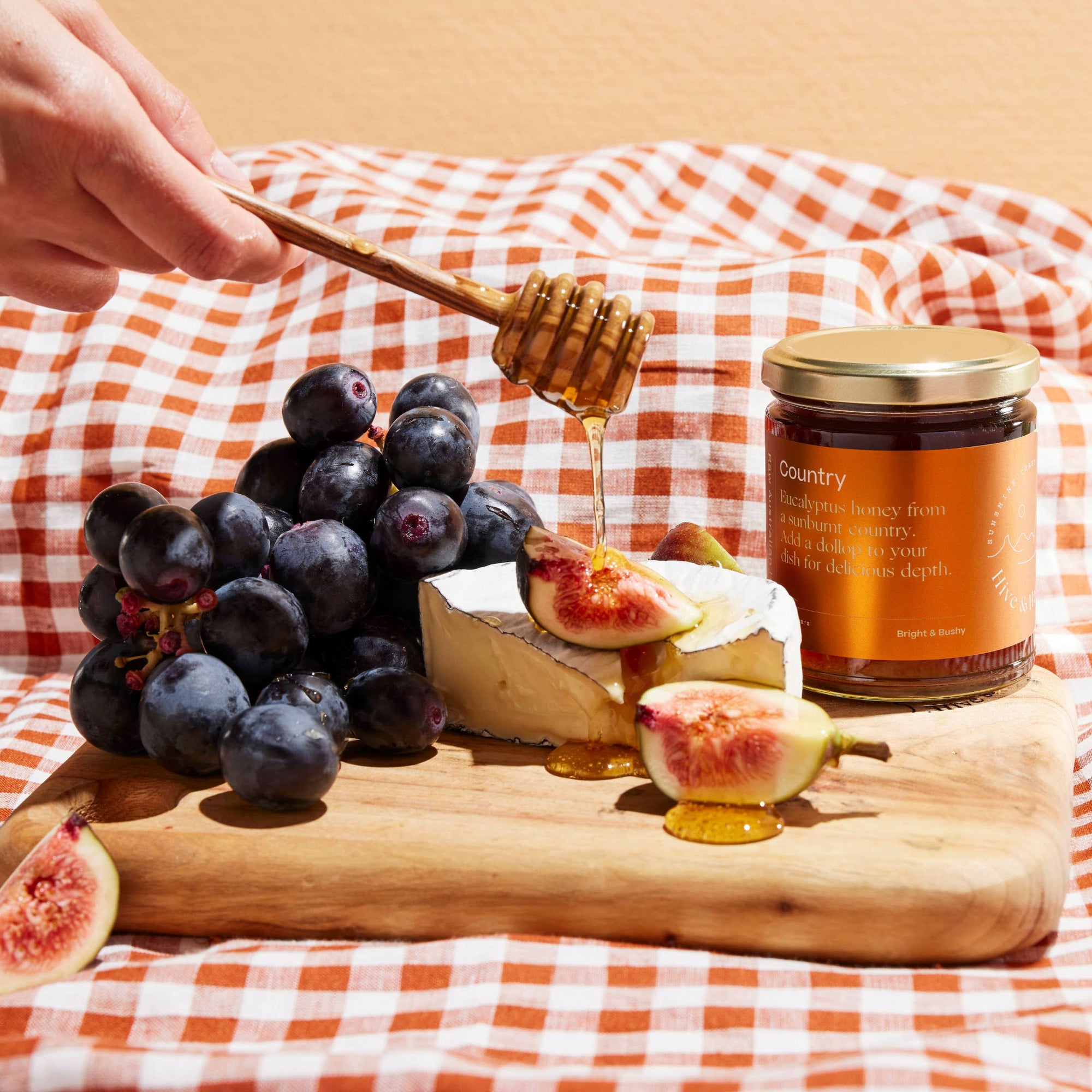 Honey being drizzled over fruit and cheese on a wooden board with a jar of honey and plaid cloth in the background.