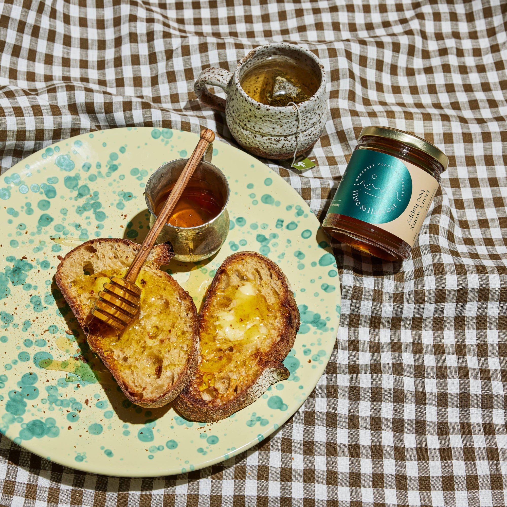 Two pieces of toast with honey on a speckled plate, accompanied by a jar of jam and two cups on a checkered tablecloth.