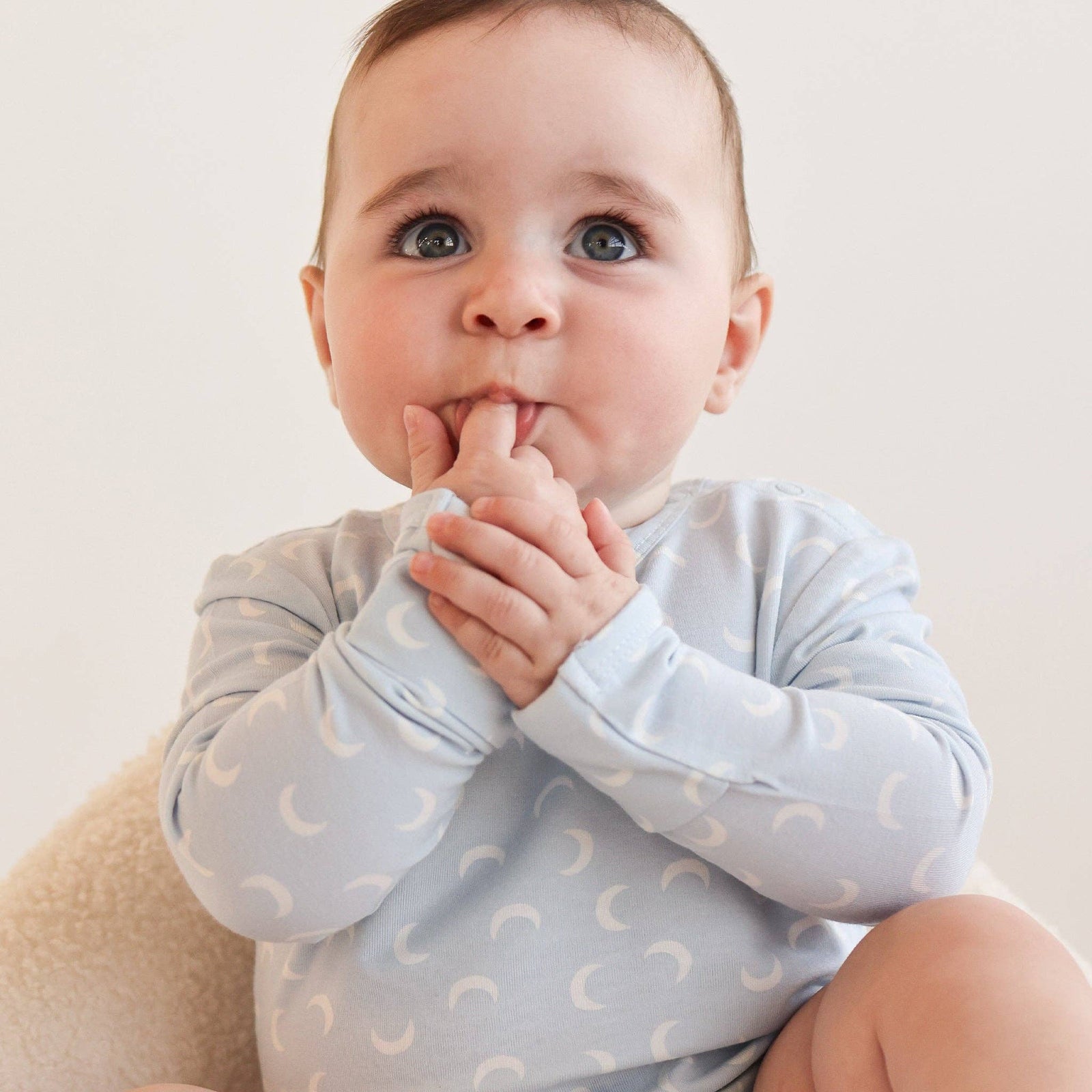 Baby sitting on a beige chair wearing a light blue onesie with white patterns.