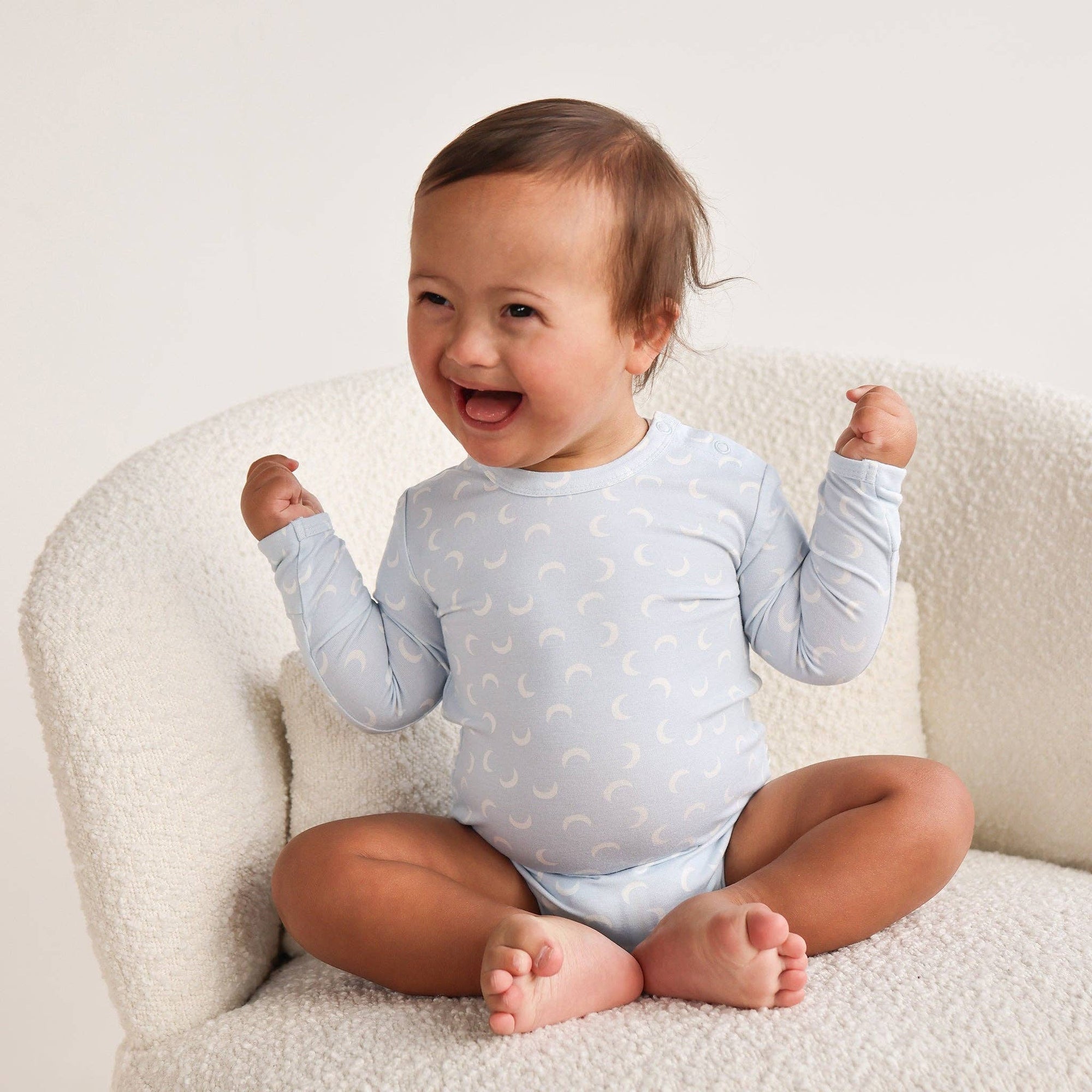 Baby sitting on a white chair wearing a light blue onesie with a pattern.