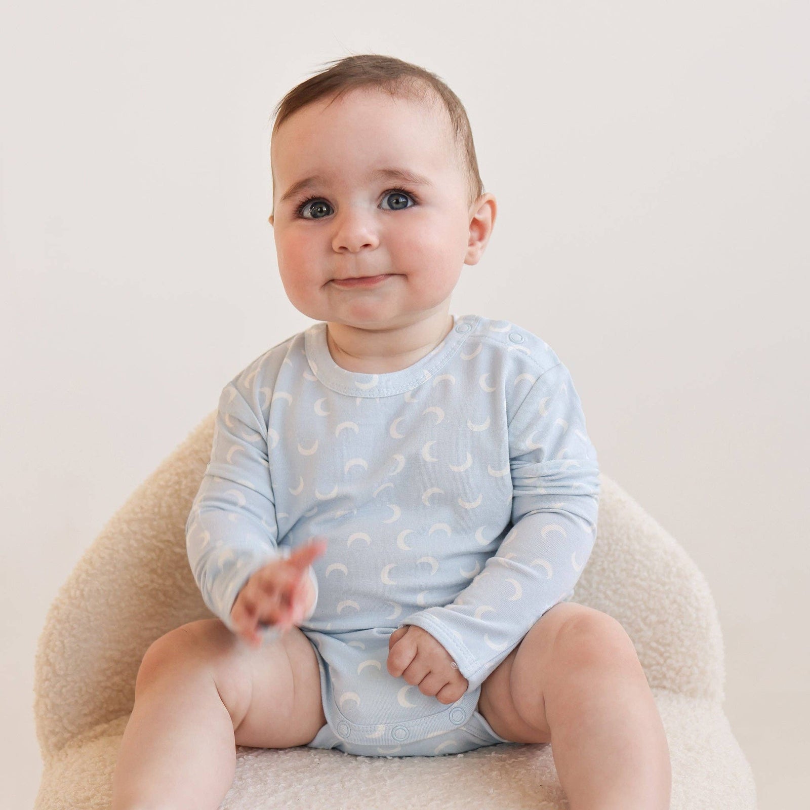 Baby sitting on a beige chair wearing a light blue onesie with white patterns.