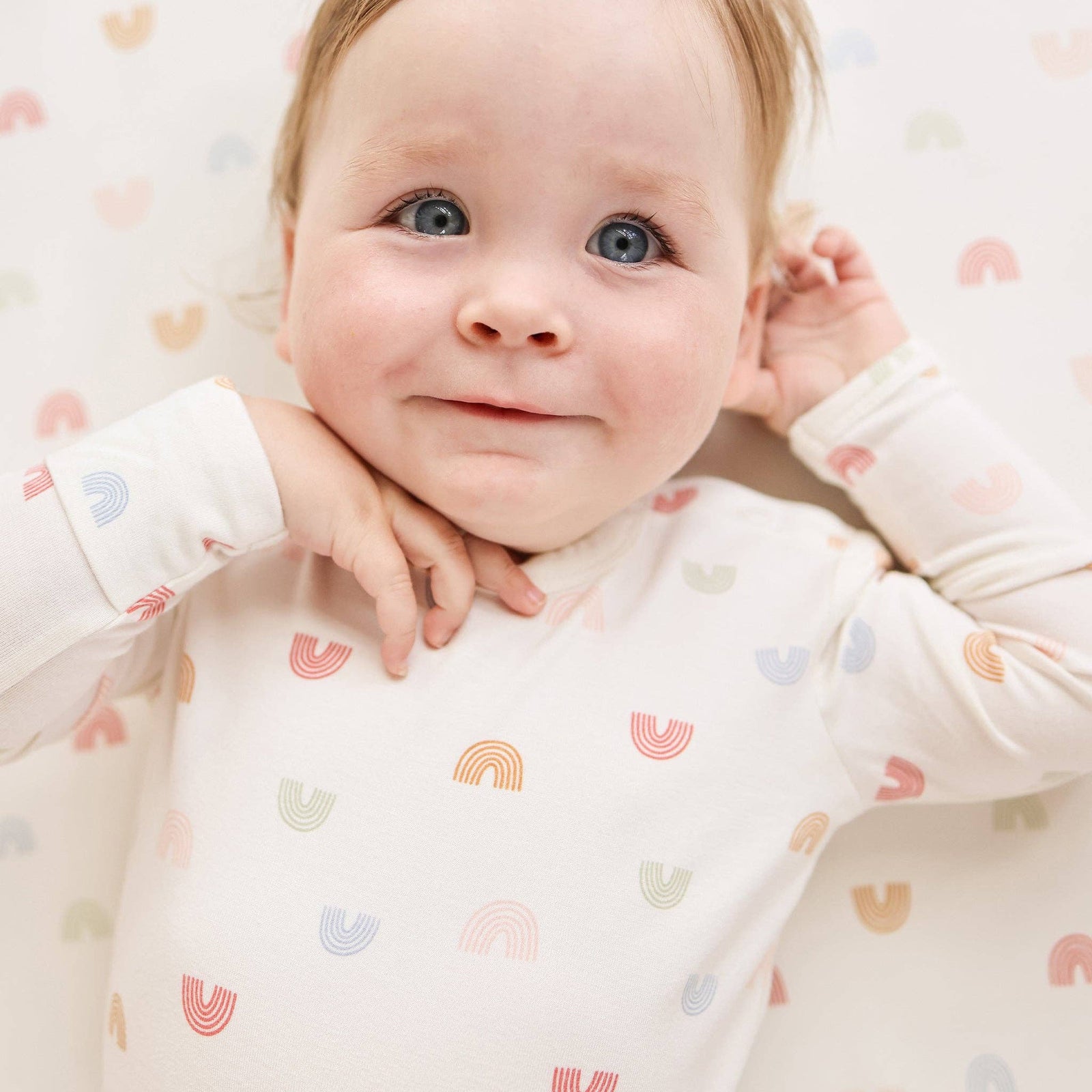 Baby wearing a white onesie with rainbow patterns sitting on a beige couch.