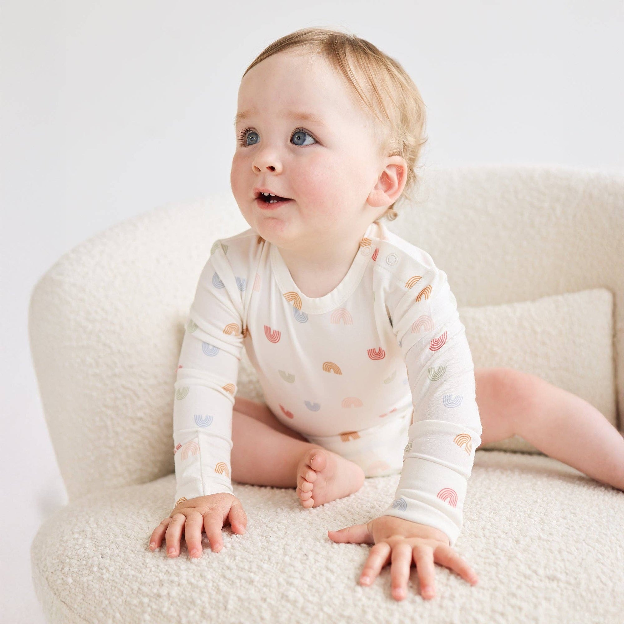 Baby sitting on a white couch wearing a white onesie with colorful patterns.
