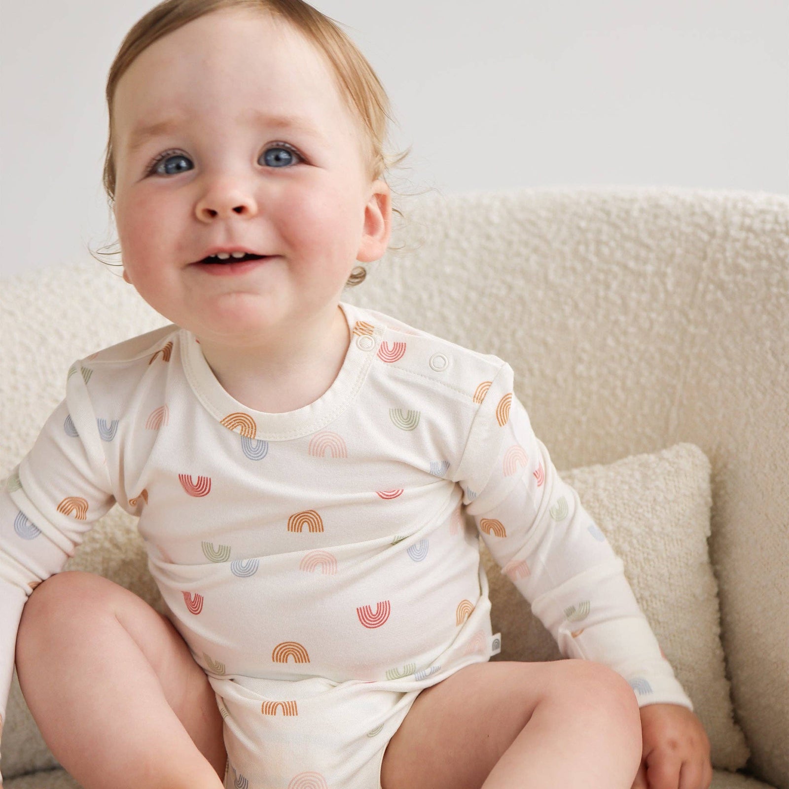 Baby wearing a white onesie with rainbow patterns sitting on a beige couch.