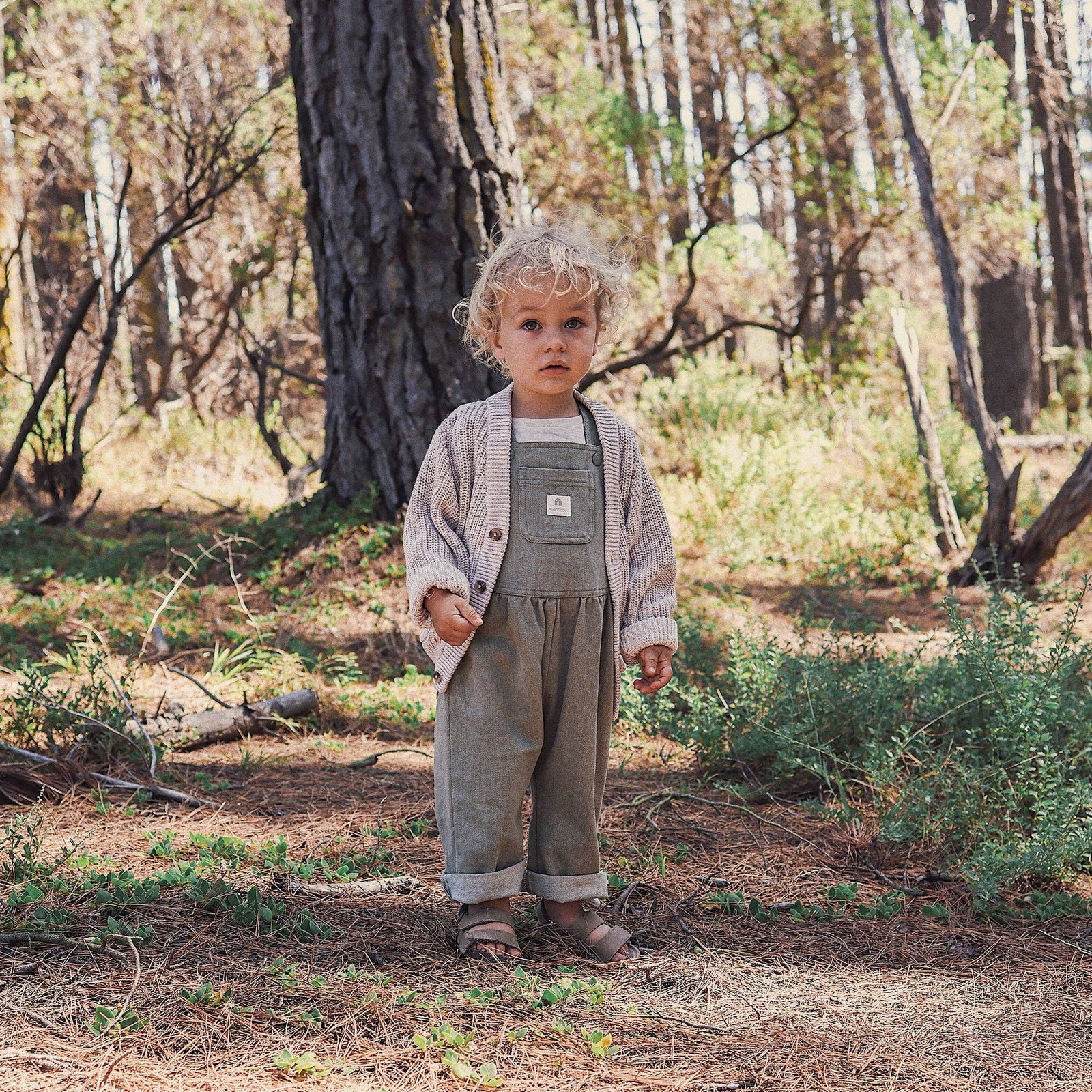 Child wearing a beige cardigan over a green outfit in a forest setting