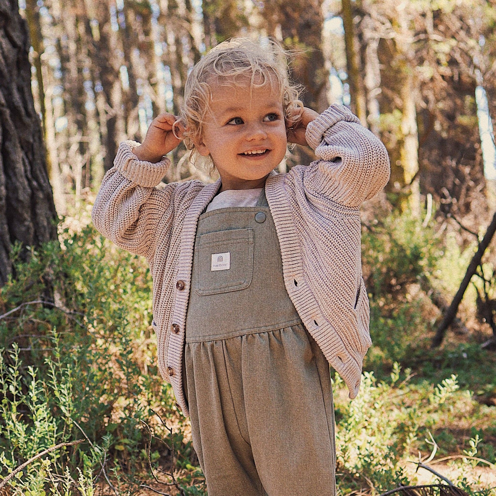 Child wearing a beige cardigan over a green outfit in a forest setting