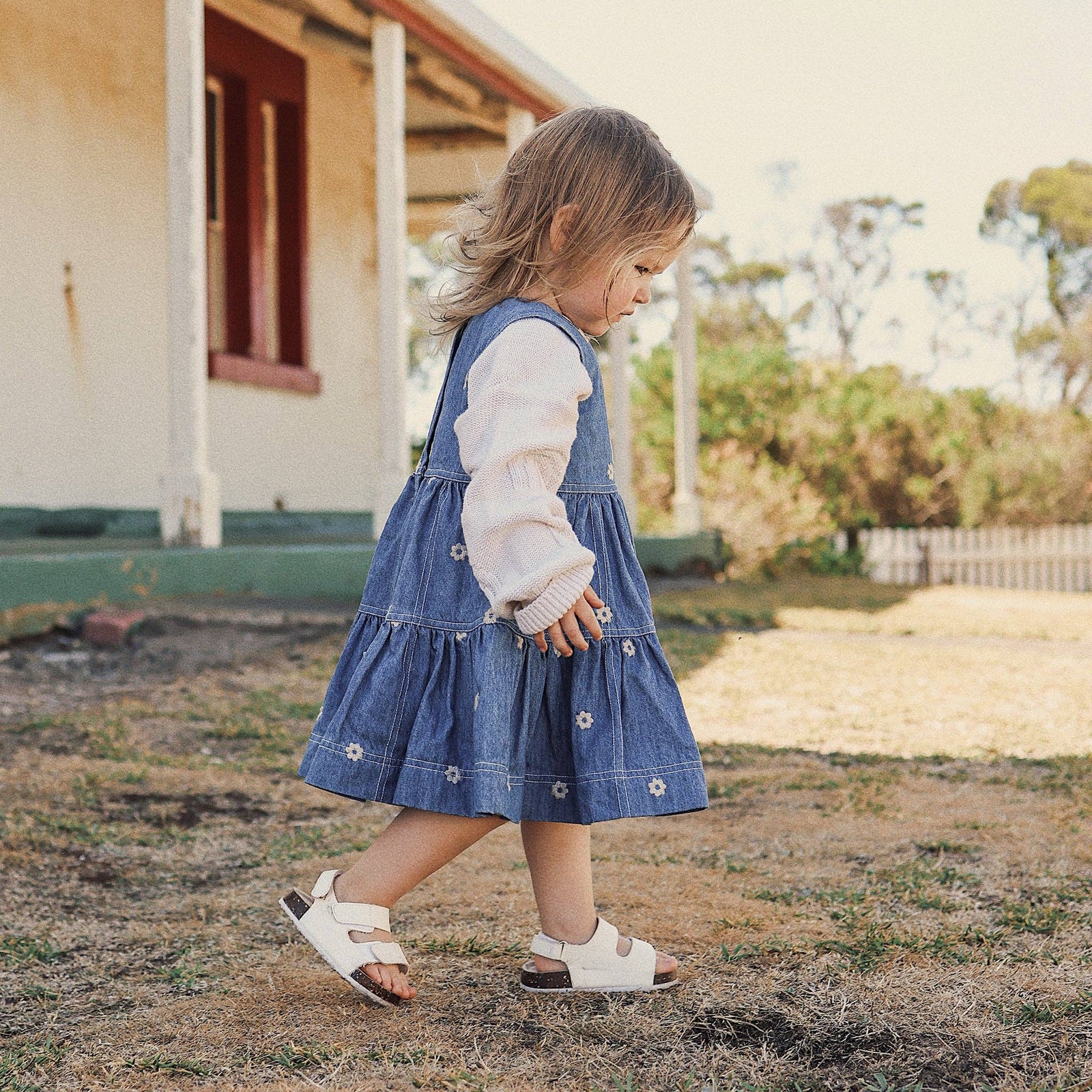 Young girl in a denim dress standing in front of a house.