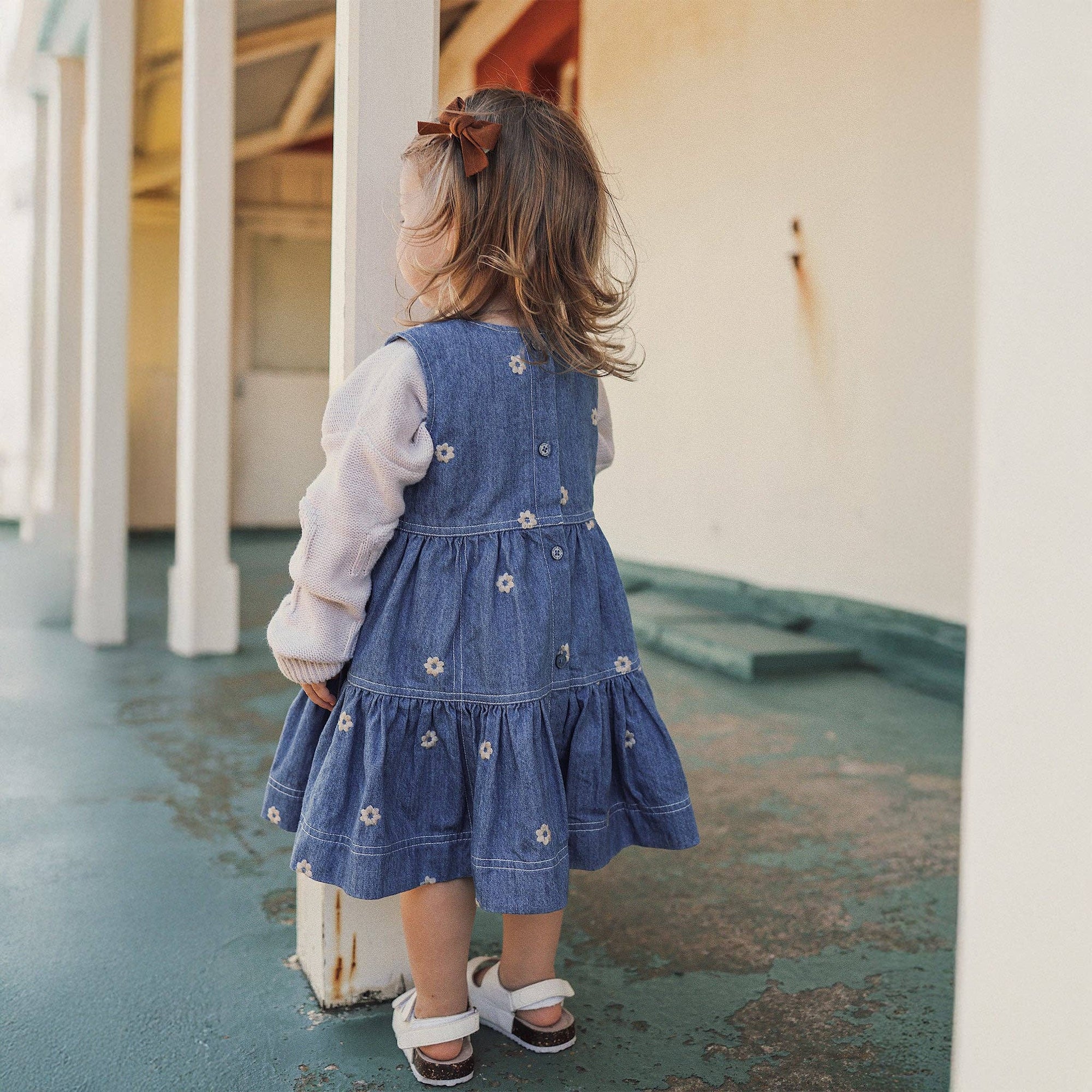 Child wearing a denim dress with floral embellishments, standing on a concrete floor.