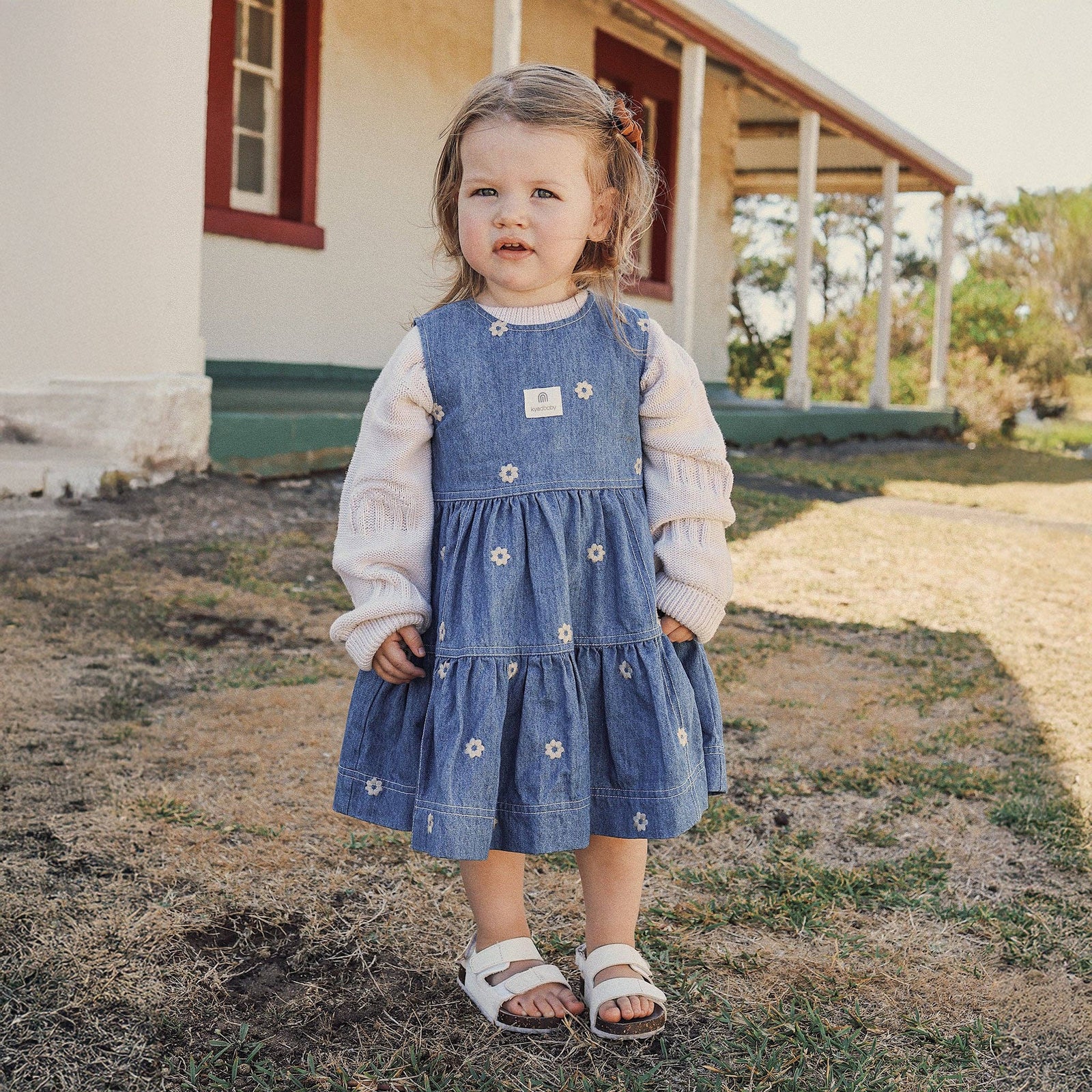 Young girl in a denim dress standing in front of a house.
