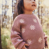 Child wearing a pink sweater with white floral patterns in a forest setting