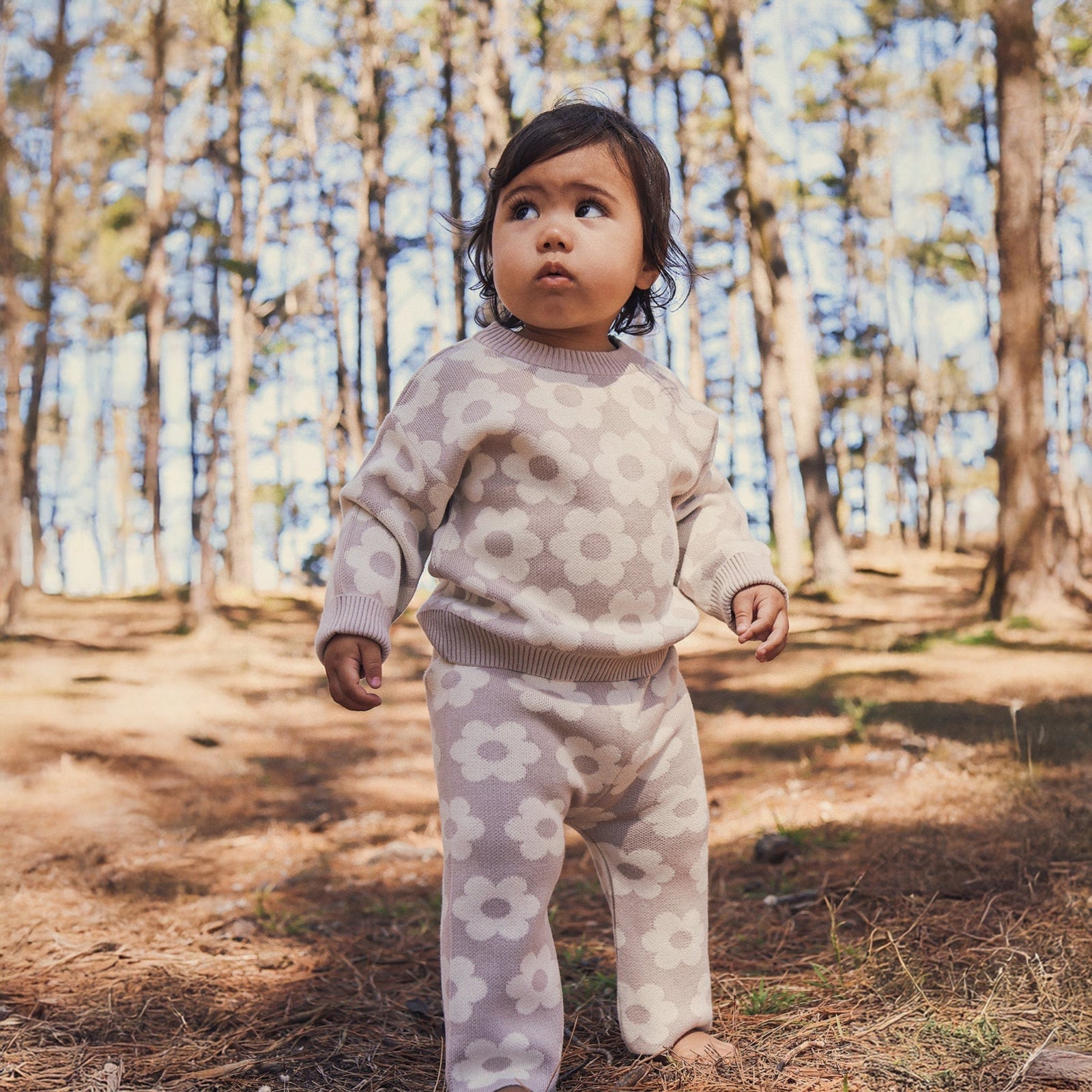 Child in a floral outfit holding a pine cone in a forest setting