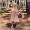 Child in a pink dress walking through a forest with a woven bag.