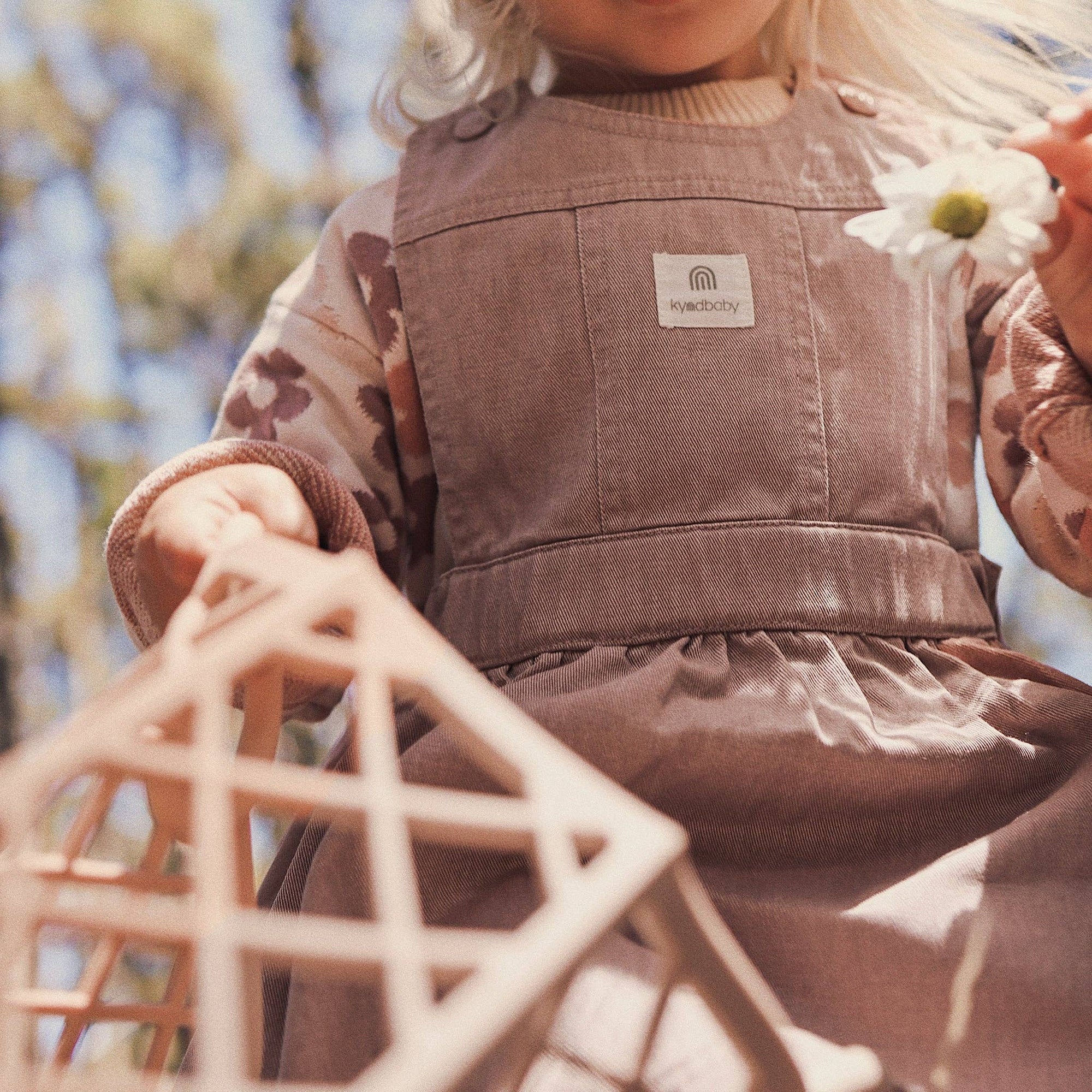 Child wearing a brown corduroy dress with a brand logo, standing outdoors with blurred background