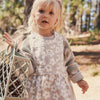 Child wearing a floral dress with a knit sweater, standing outdoors in a natural setting.