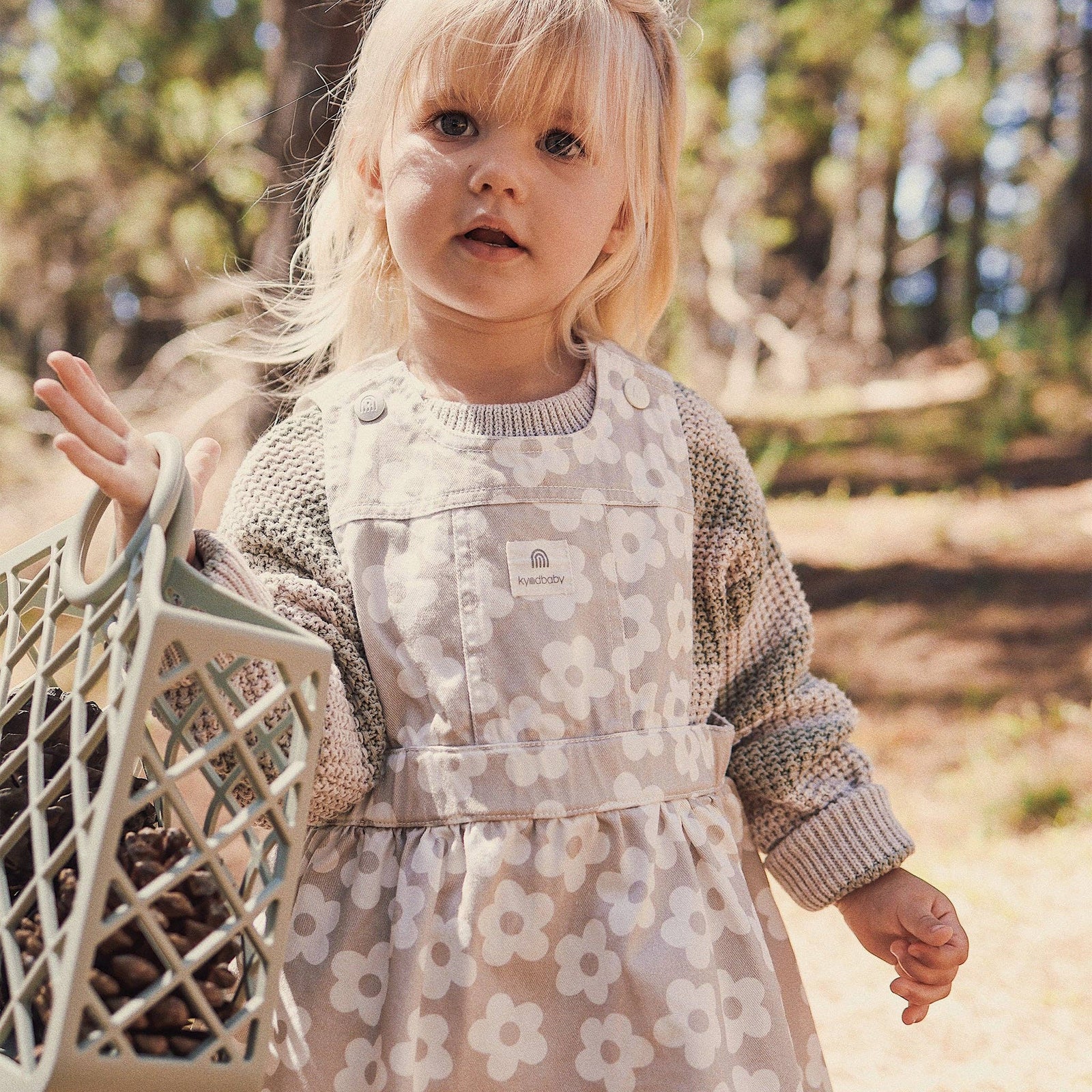 Child wearing a floral dress with a knit sweater, standing outdoors in a natural setting.