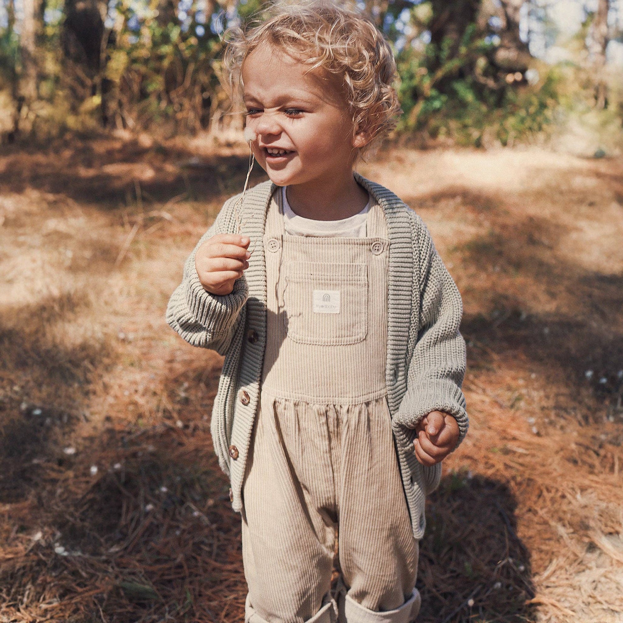 Child wearing beige overalls and a gray cardigan standing in a natural setting