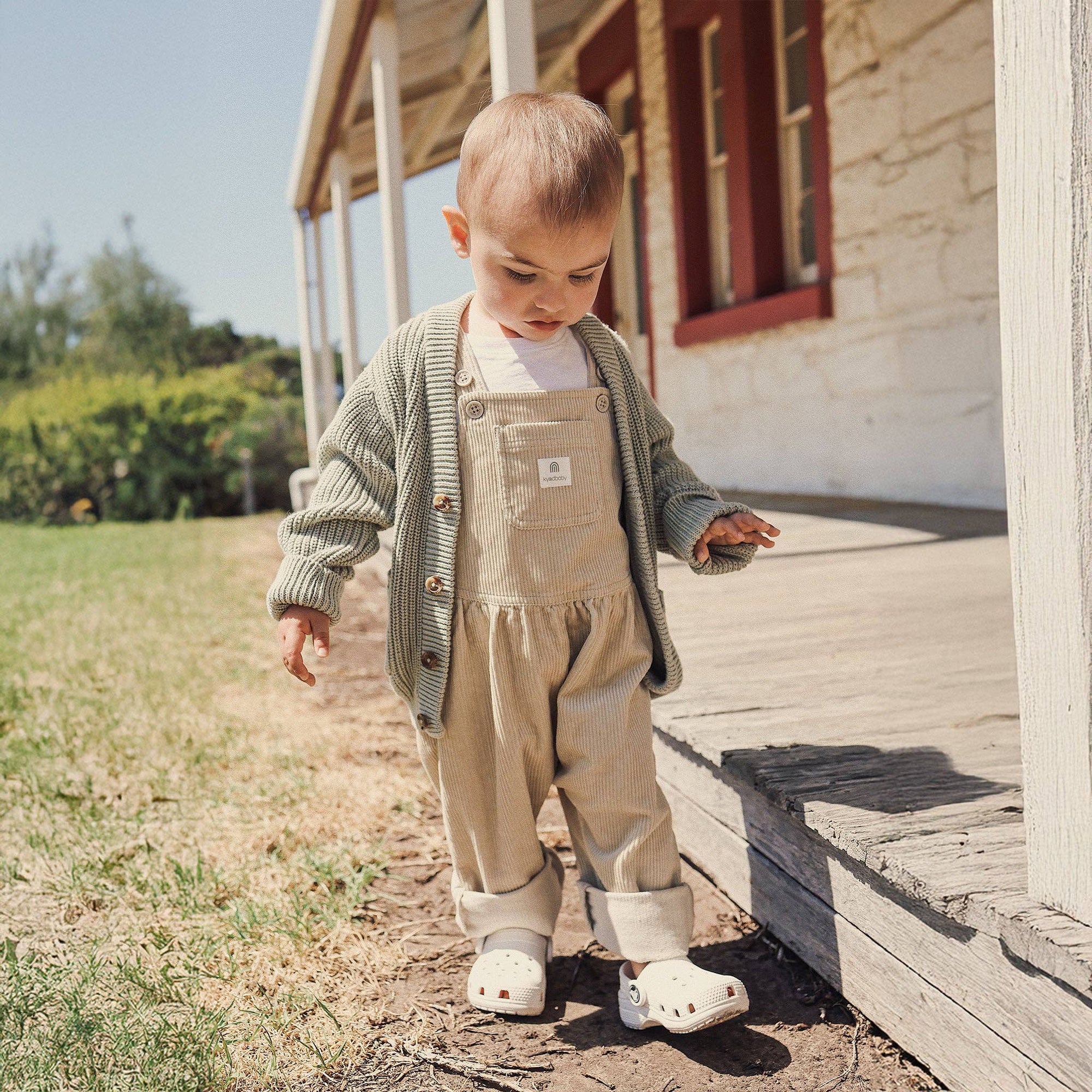 Child in beige overalls and green cardigan standing on a wooden porch.