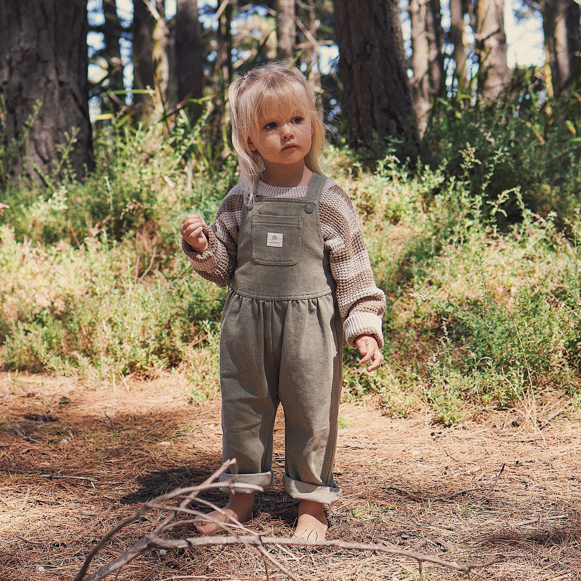 Child in a forest wearing a green overalls and patterned shirt.