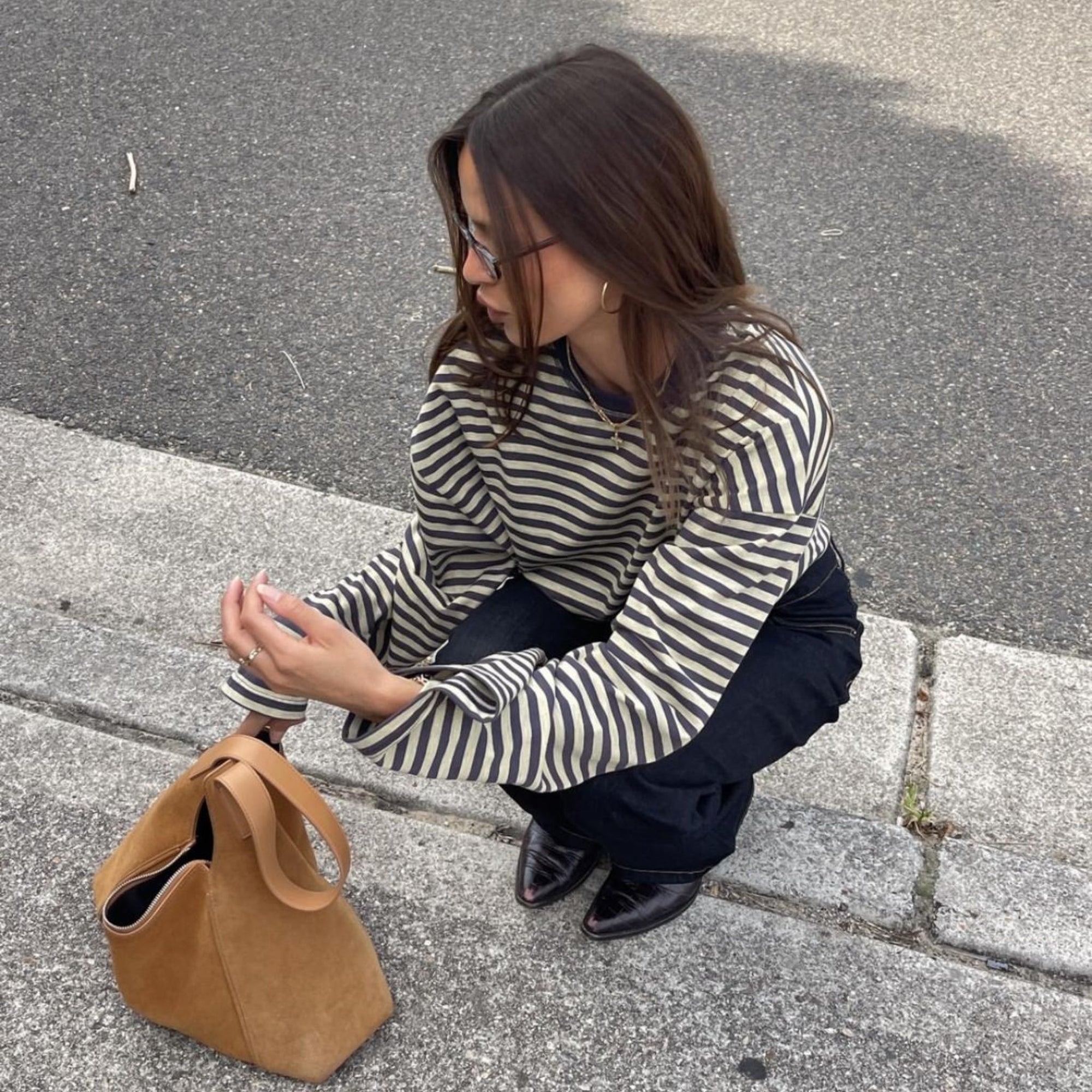 Woman in striped sweater and dark pants squatting on a sidewalk with a brown bag.