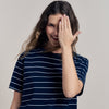 Woman wearing a navy striped shirt and beige pants, covering her face with one hand on a plain background